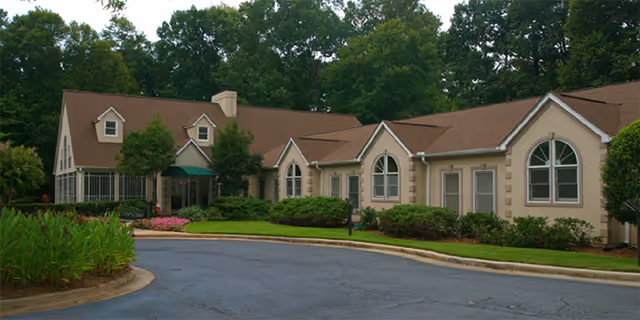 Front exterior of a one-story beige residential-style building with arched windows, a curved driveway, and landscaped greenery.