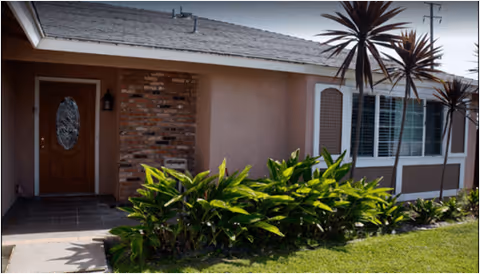 Exterior view of a single-story residential building with a wooden front door featuring an oval glass panel, brick and stucco walls, white-framed windows with shutters, and a garden with green plants and grass in front.