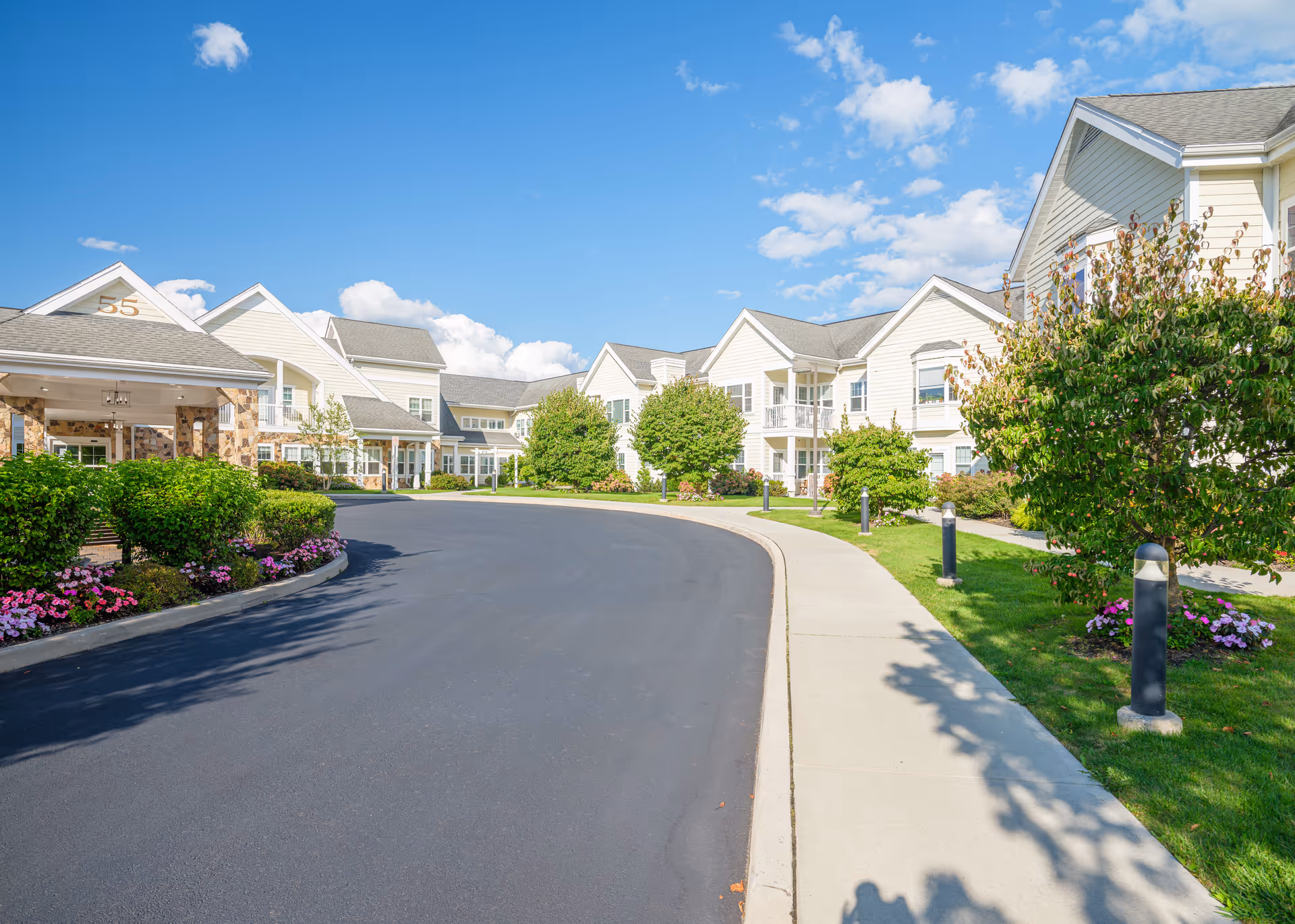 Exterior view of a senior living facility with a curved driveway, well-maintained landscaping including bushes and flowers, and multiple two-story buildings under a blue sky with scattered clouds.