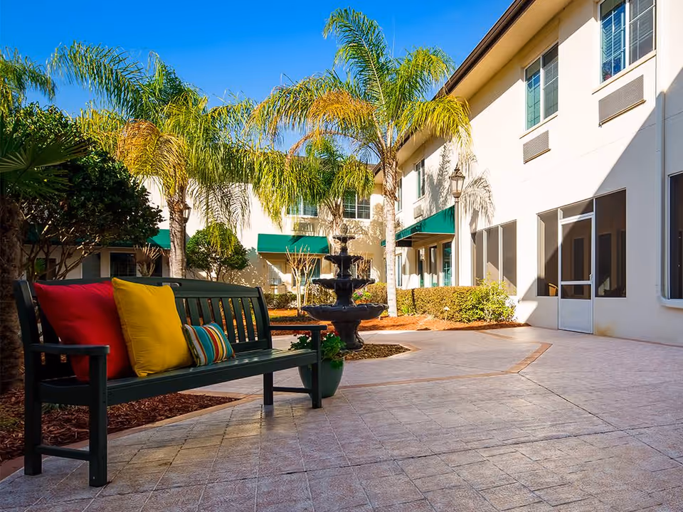 Outdoor courtyard area at The Windsor Of Ocala featuring a green bench with colorful cushions, palm trees, a multi-tiered water fountain, and a building with green awnings and large windows under a clear blue sky.