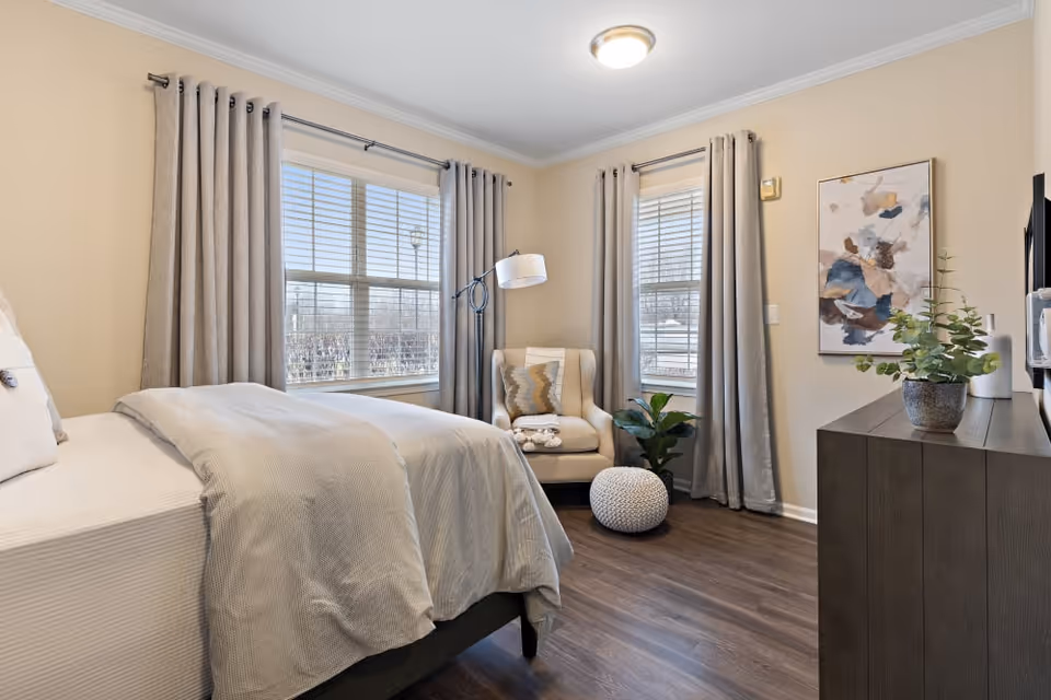 A cozy bedroom with a neatly made bed covered in light gray bedding, two large windows with beige curtains, a beige armchair with a decorative pillow and folded towels, a round knitted pouf, a dark wooden dresser with a potted plant, and abstract wall art. The room has wood flooring and soft lighting.