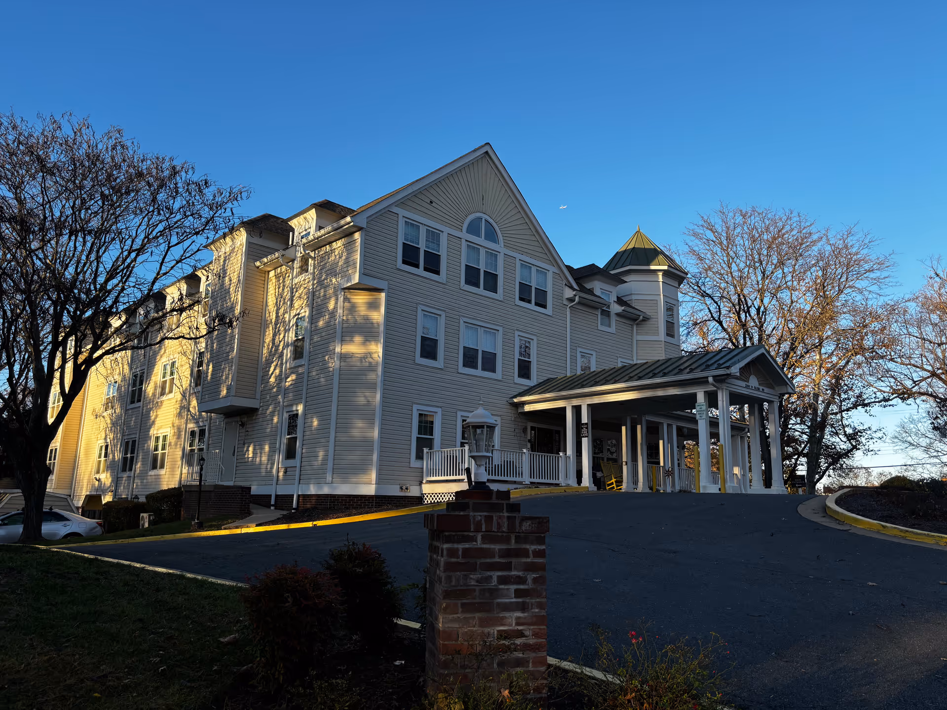 Front exterior view of a multi-story beige senior living building with a covered entrance, driveway, and surrounding trees at sunset.