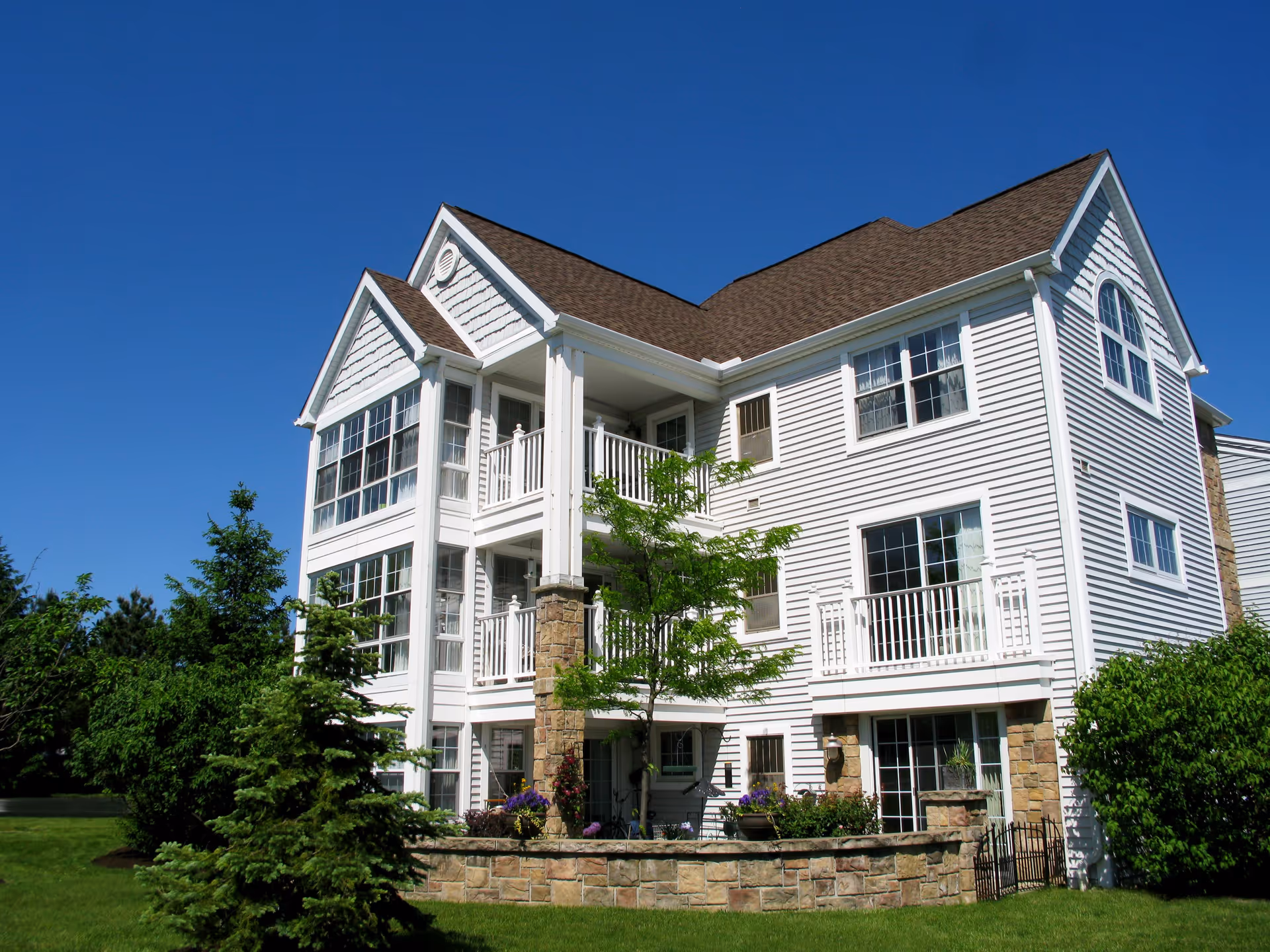 Exterior view of a multi-story white residential building with large windows, balconies, and a stone base, surrounded by green trees and grass under a clear blue sky.
