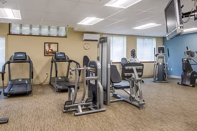 A fitness room with exercise equipment including two treadmills, weight machines, and stationary bikes. The room has beige walls, a blue accent wall, carpeted floor, and windows with blinds letting in natural light. A wall clock and a framed picture are visible on the wall.