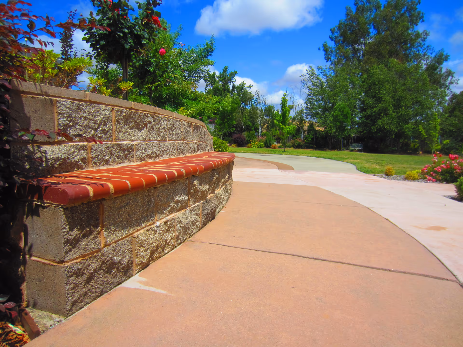 Curved outdoor pathway with a low stone wall topped with red bricks on the left side, surrounded by green trees, bushes, and flowers under a partly cloudy blue sky.