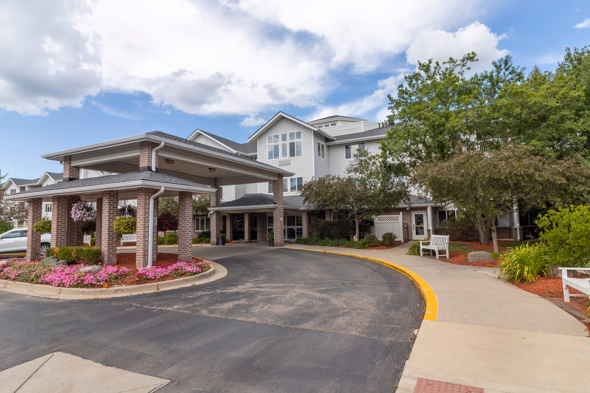 Exterior view of StoryPoint East Lansing senior living facility showing a covered entrance with brick pillars, landscaped flower beds with pink flowers, trees, benches, and a multi-story building under a partly cloudy sky.