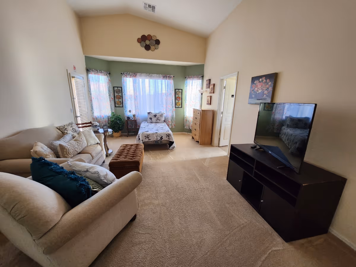 A cozy living area with a beige sofa adorned with multiple pillows, a brown ottoman, and a TV on a dark wooden stand. In the background, there is a single bed with floral bedding positioned in front of three windows with floral curtains. A wooden dresser and a floor lamp are also visible near the bed. The walls are painted in light beige and green tones, and the room is carpeted.