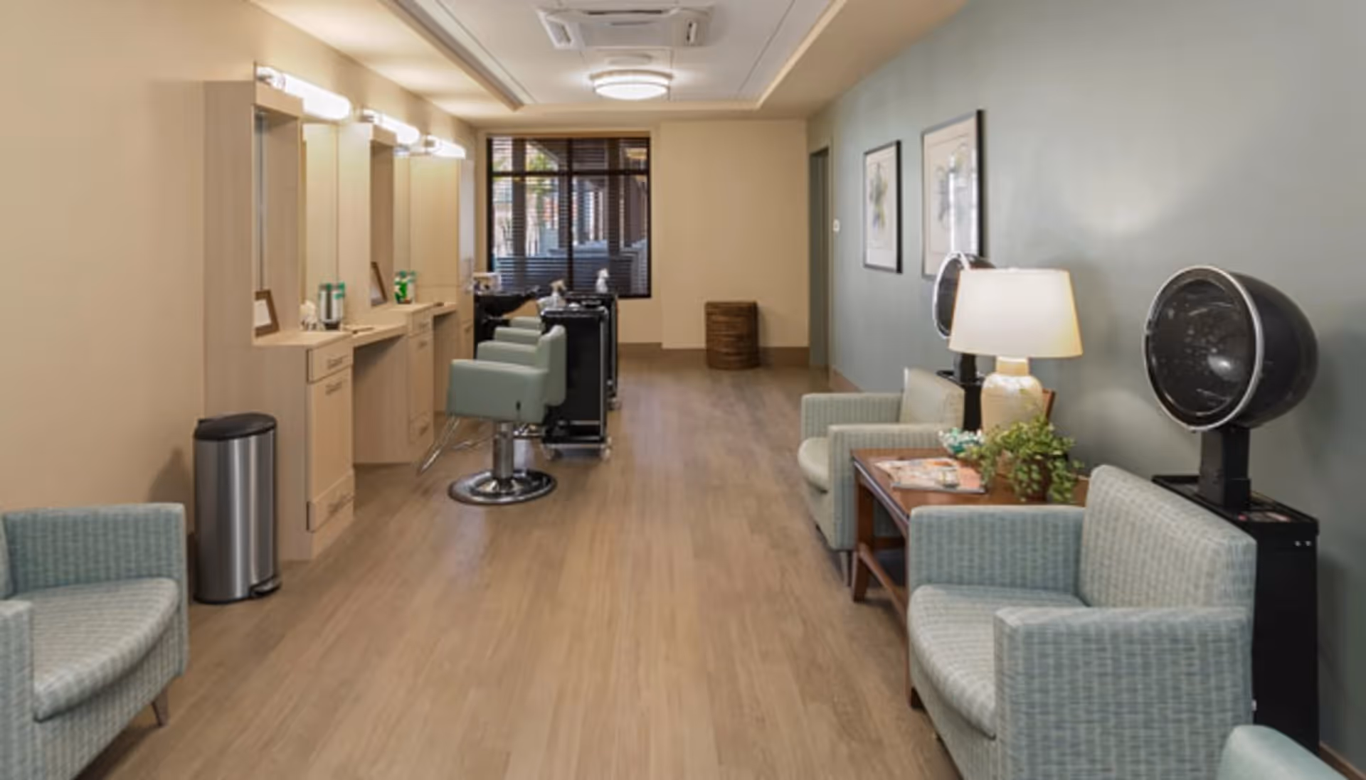 Interior view of a salon area in a senior living facility with salon chairs and mirrors on the left side, and a seating area with armchairs, a table with a lamp and plants, and hair dryers on the right side.