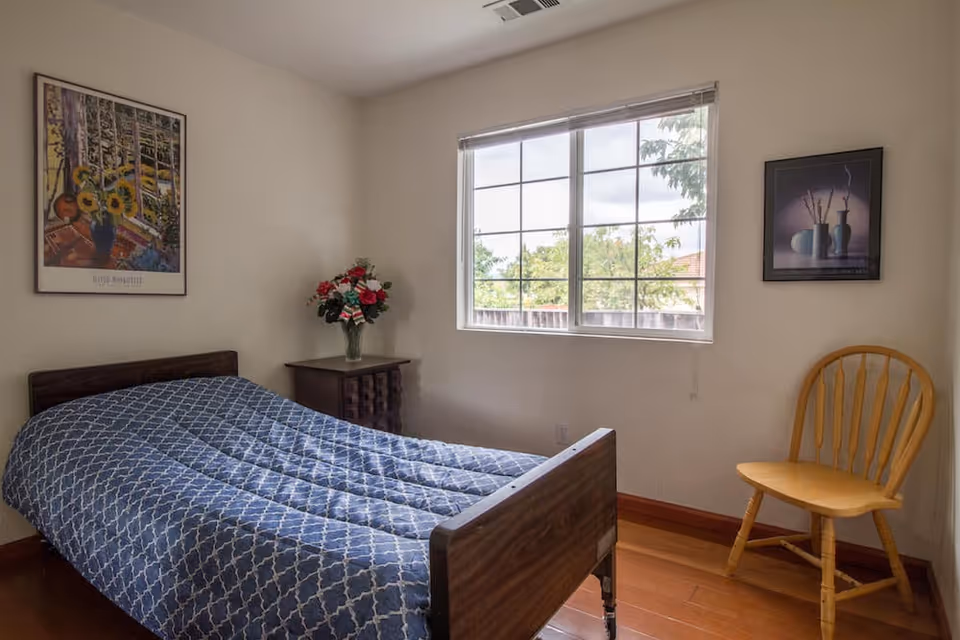 A simple bedroom with a single bed covered in a blue patterned quilt, a wooden nightstand with a vase of flowers, a wooden chair, two framed pictures on the walls, and a window showing trees outside.