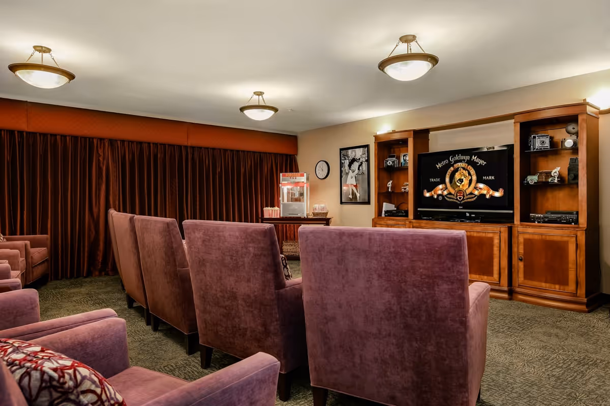 A cozy senior living facility media room with multiple plush purple armchairs arranged in rows facing a large wooden entertainment center with a television displaying the Metro Goldwyn Mayer logo. The room has warm lighting, dark curtains covering the windows, a popcorn machine on a side table, and framed black and white photos on the walls.