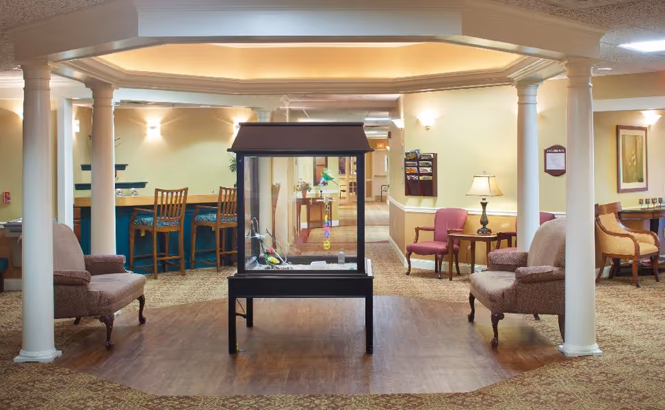 A well-lit interior common area in a senior living facility featuring a central glass display case with decorative items inside. The room has beige walls, wooden flooring in the center, and carpeted areas around. There are several upholstered chairs and wooden chairs arranged around the space, with a small table and lamp on the right side. The ceiling has recessed lighting and decorative molding supported by white columns.