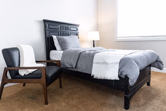 Single bed with gray bedding, a bedside lamp, and a wooden armchair in a bright bedroom.