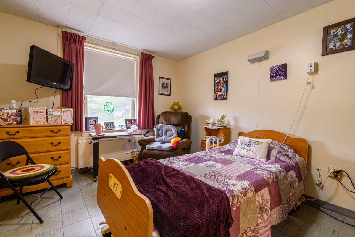 A cozy bedroom in a rehabilitation and nursing center featuring a single bed with a purple and white patchwork quilt and a decorative pillow. Next to the bed is a small wooden nightstand with various items on it. A comfortable brown recliner chair with a cushion is positioned near a window with red curtains and a white roller shade. A wooden dresser with greeting cards and a wall-mounted TV are also visible. The walls are decorated with framed photos and artwork.
