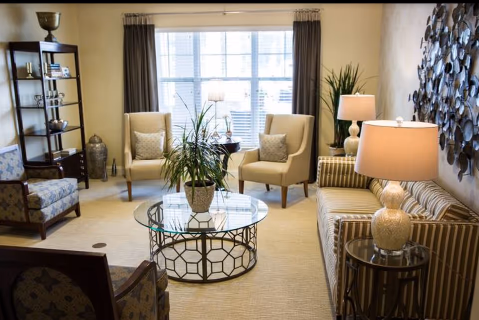 A cozy living room with a large window covered by dark curtains, two beige armchairs with patterned cushions, a striped sofa with a table lamp on a round side table, a glass coffee table with a potted plant, a tall bookshelf with decorative items, and a large metal wall art piece.