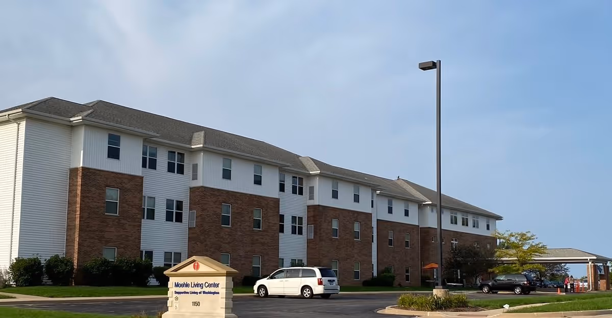 Exterior view of a three-story senior living facility building with a combination of brick and white siding. There is a parking lot in front with several cars parked and a sign that reads 'Moehle Living Center Supportive Living of Washington 1150'. The sky is clear with some light clouds.