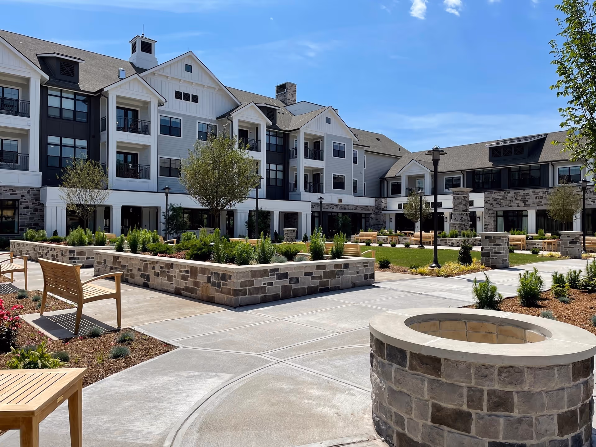 Sunny courtyard with stone planters, wooden benches and a circular stone fire pit in front of a multi-story senior living building.
