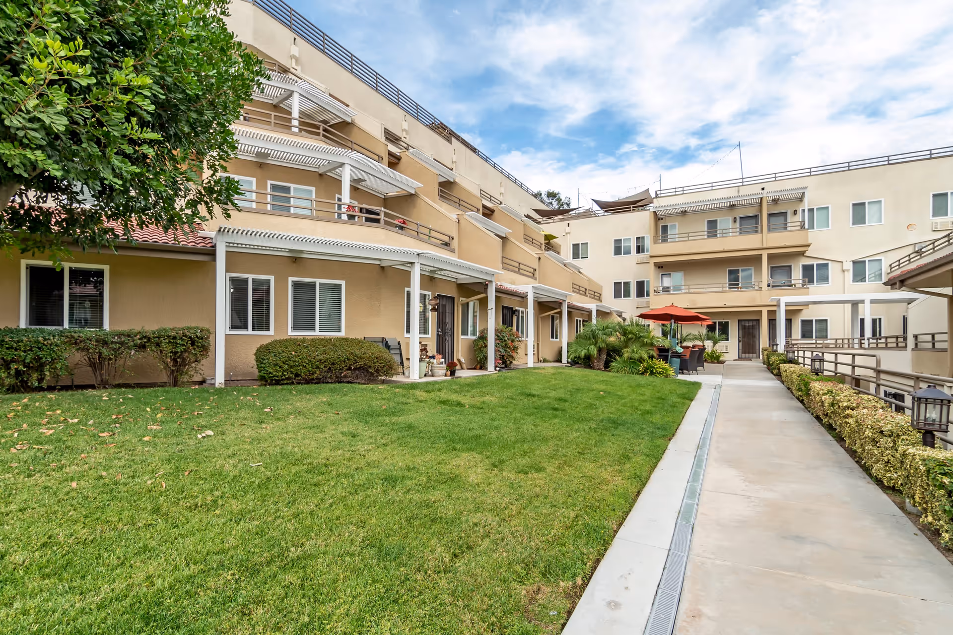 Outdoor courtyard area of a multi-story retirement community building with beige walls, balconies, green lawn, shrubs, and a paved walkway. There are patio tables with umbrellas and chairs near the building.