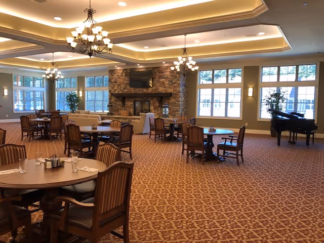 Spacious assisted-living dining and lounge area with round tables and chairs, chandeliers, a stone fireplace, large windows, and a grand piano.