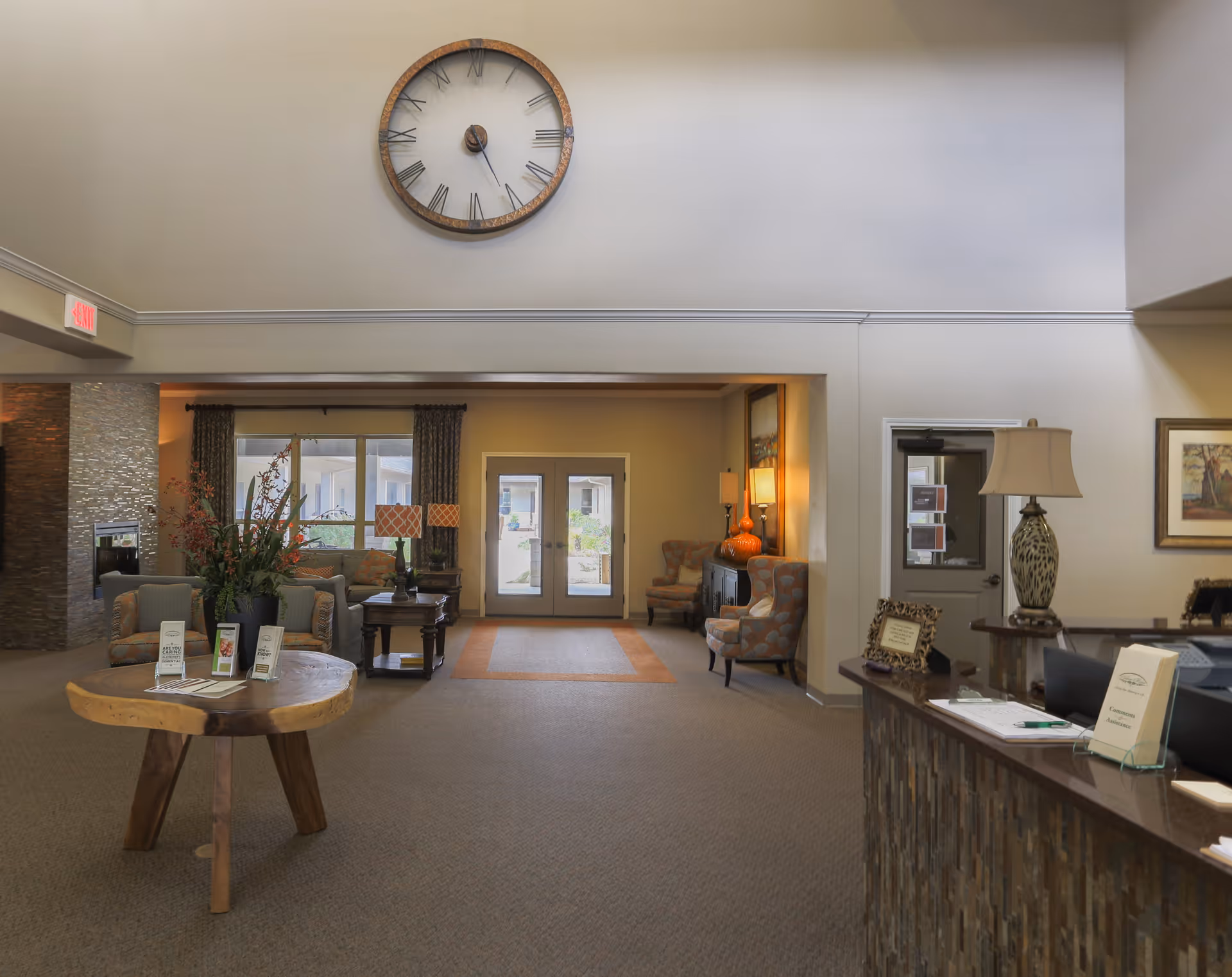 A spacious assisted living facility lobby with a large round wall clock above the entrance. The room features a wooden table with brochures, comfortable armchairs, a fireplace, and a reception desk with a lamp and informational materials. Double glass doors lead outside, and the area is warmly lit with lamps and natural light from windows.