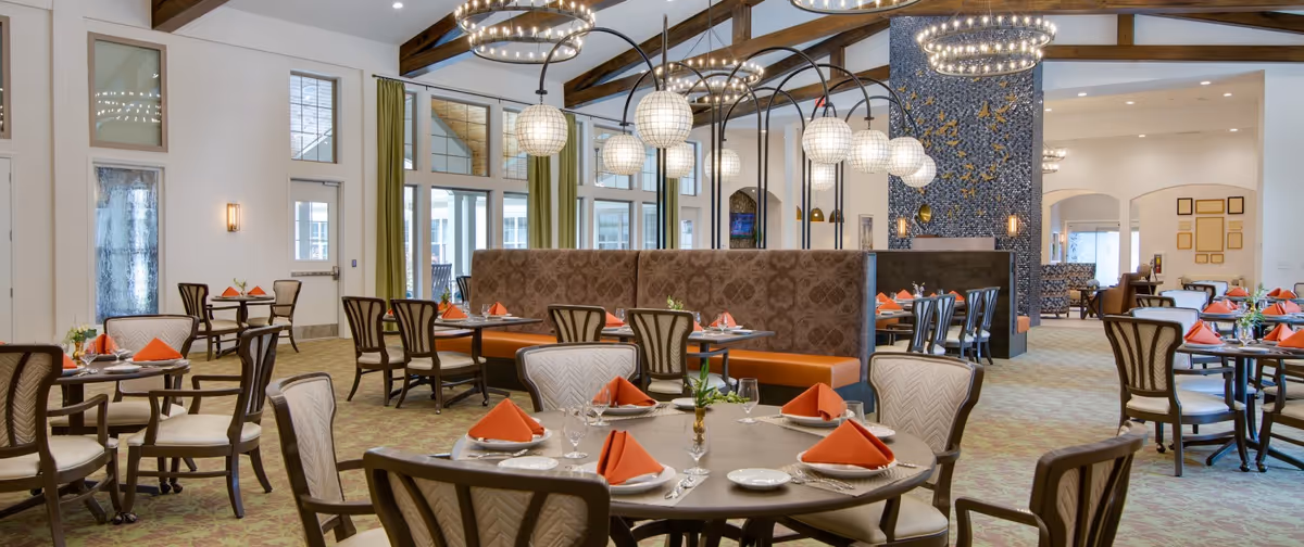 Spacious, well-lit dining room with round tables set with orange napkins, upholstered chairs, chandeliers, and high beamed ceilings.