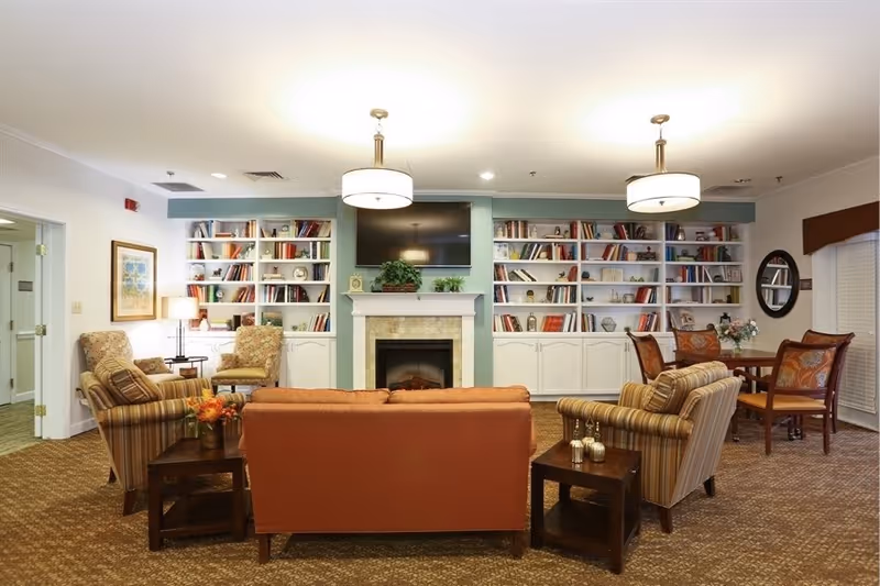 A cozy living room area with a burnt orange sofa facing a fireplace with a mounted TV above it. There are built-in white bookshelves filled with books and decorative items on either side of the fireplace. Two striped armchairs flank the sofa, and a wooden side table with small decorative pumpkins is beside one armchair. In the background, there is a small round dining table with four chairs, a round mirror on the wall, and window blinds. The room is warmly lit with ceiling lights and a table lamp.