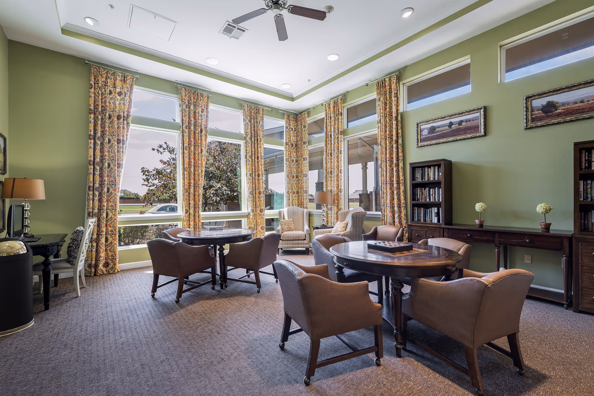 Bright common lounge with round tables and upholstered chairs in front of tall windows and patterned curtains.