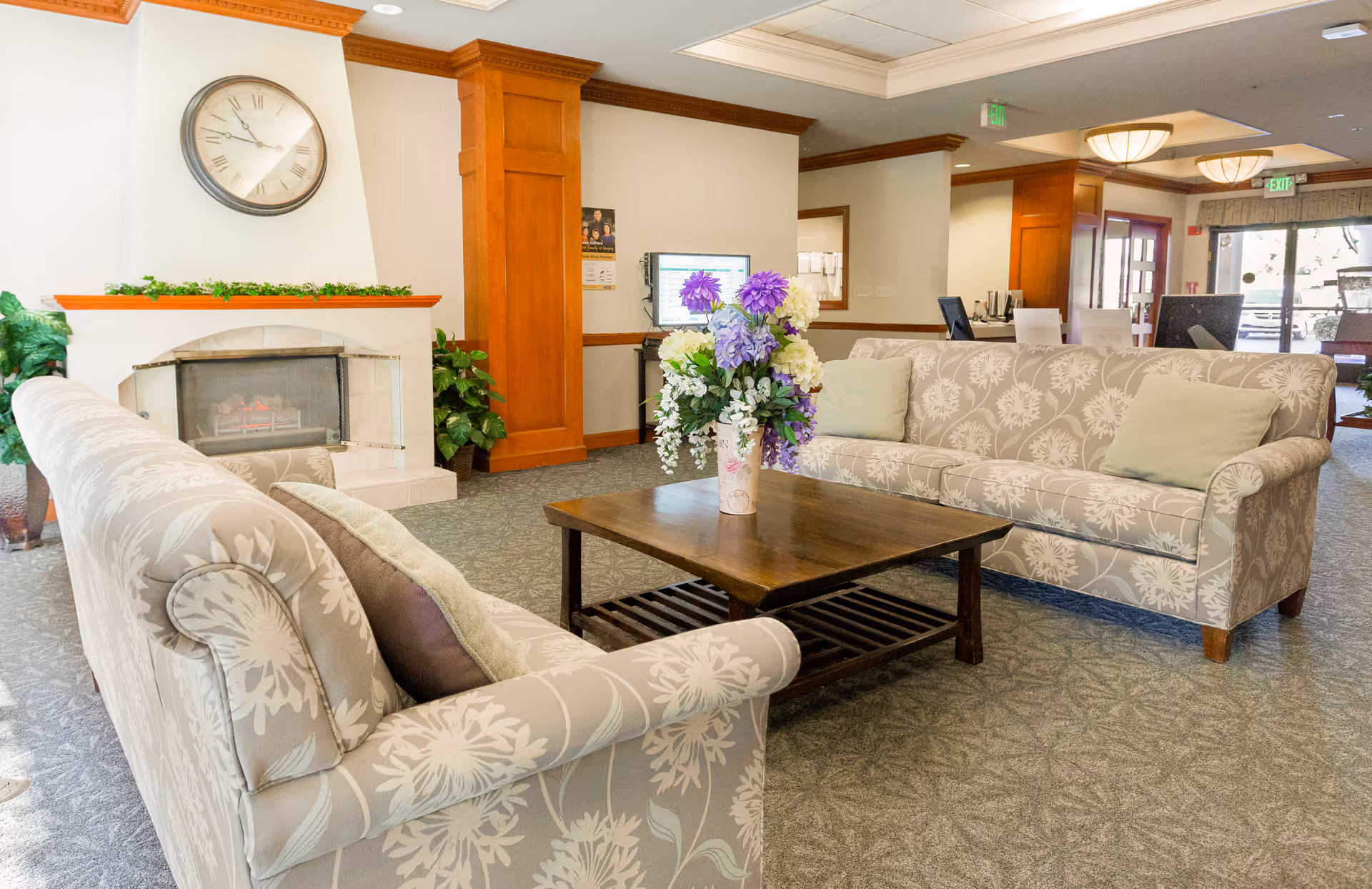 Bright seating area with two patterned sofas facing a wooden coffee table topped with a floral arrangement, a fireplace and clock on the wall, and a reception desk in the background.