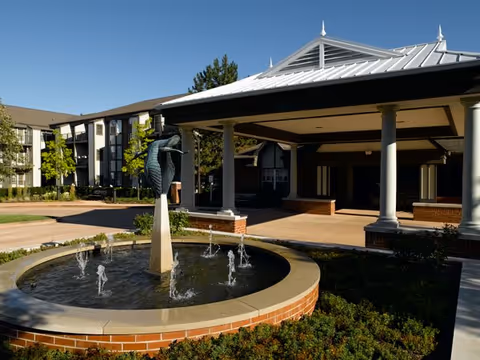 Outdoor view of Atria Kinghaven featuring a circular water fountain with a bird sculpture in the center, surrounded by greenery and a covered entrance with white columns and a metal roof. Residential building with multiple windows is visible in the background under a clear blue sky.