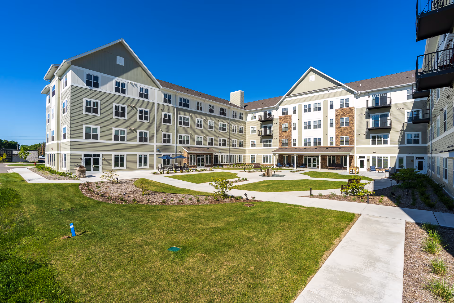 Large four-story senior living building surrounding a landscaped courtyard with walkways and seating under a clear blue sky.