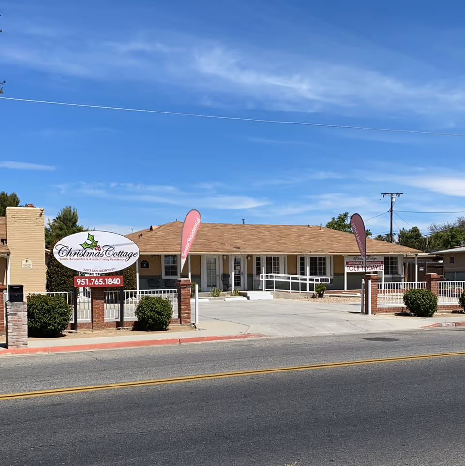 Exterior view of a single-story senior living facility named Christmas Cottage with a beige roof and white walls. The building is surrounded by a low brick and metal fence with bushes in front. There are two vertical flags on either side of the entrance and a sign displaying the facility name, address, and phone number. The sky is clear and blue.