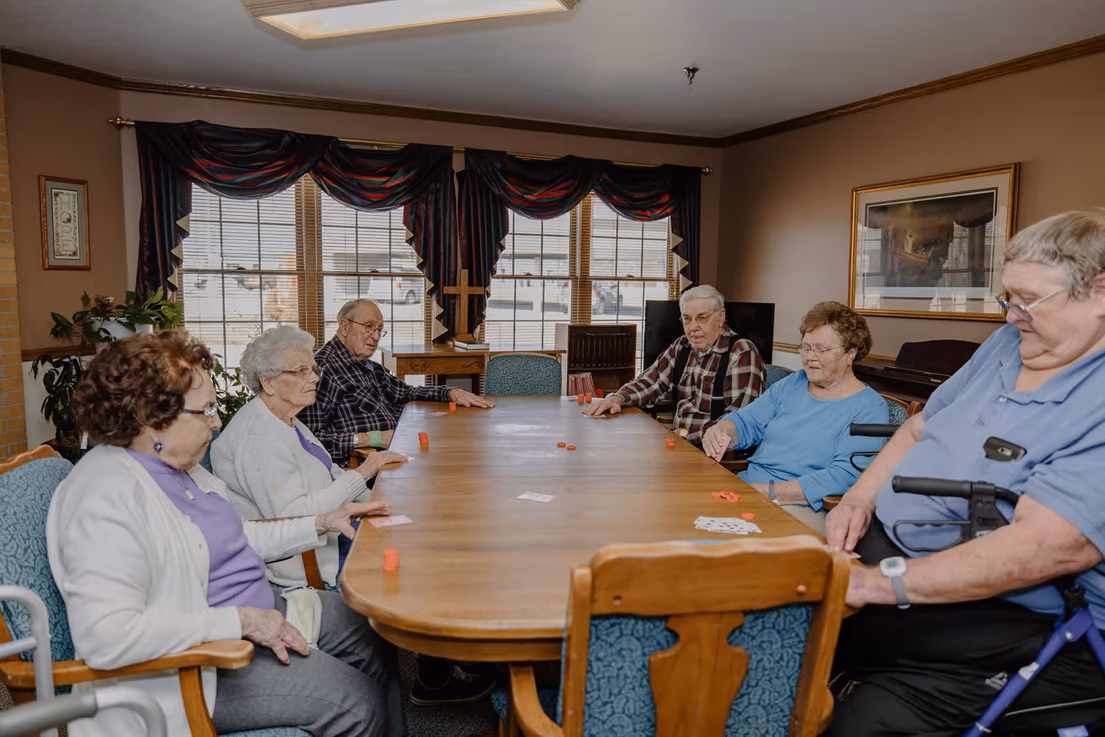 A group of six elderly people sitting around a large wooden table in a well-lit room with windows covered by dark curtains. They appear to be playing a card game with poker chips on the table. The room has beige walls, framed artwork, and a piano in the background.