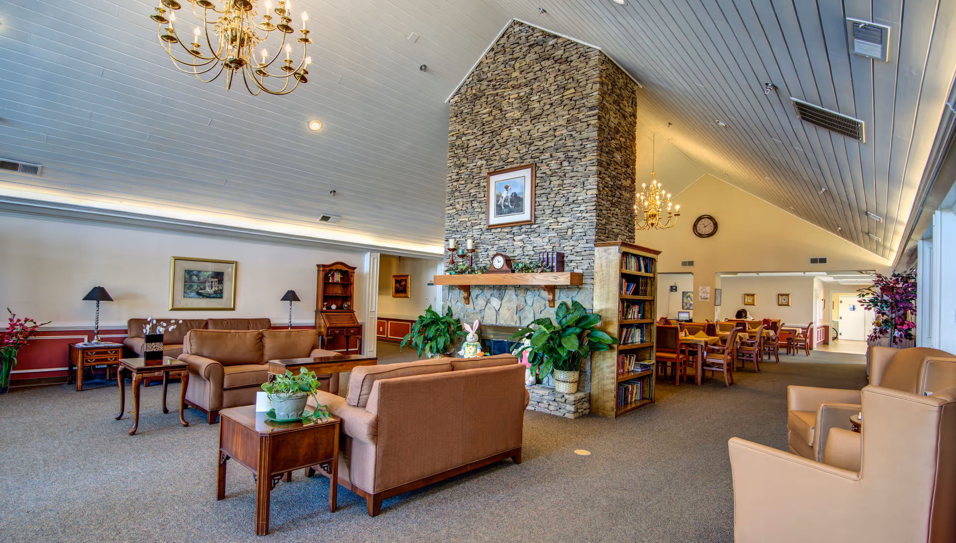 A spacious senior living facility common area with comfortable seating including sofas and armchairs arranged around a stone fireplace. The room features a high ceiling with chandeliers, framed artwork on the walls, potted plants, and a bookshelf. In the background, there is a dining area with tables and chairs.