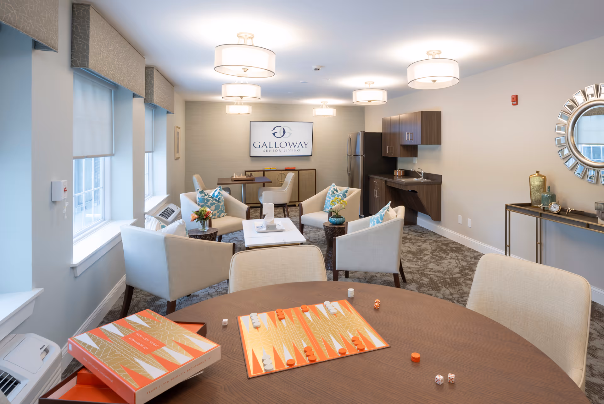 A bright and inviting common area in Galloway Senior Living featuring a round table with a backgammon game set up, several comfortable armchairs with decorative pillows arranged around a coffee table, a small kitchenette with a refrigerator and cabinets, and a wall-mounted TV displaying the Galloway Senior Living logo. The room has large windows with shades, modern ceiling lights, and a decorative mirror above a console table with vases and a clock.