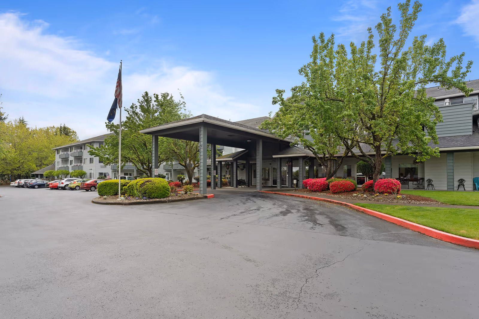 Exterior view of Parkrose Chateau Gracious Retirement Living facility showing the entrance with a covered drop-off area, surrounded by trees, bushes, and a parking lot with several cars parked.