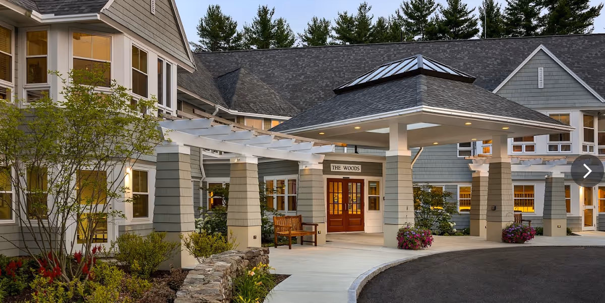 Exterior view of Riverwoods At Exeter showing the entrance area with a covered drop-off, wooden bench, landscaped garden with flowers and shrubs, and a building with multiple windows and gray siding.