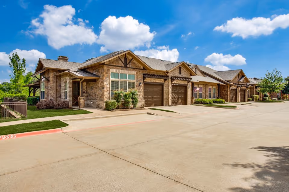 Exterior view of a row of single-story brick residential buildings with garages under a blue sky with scattered clouds. The buildings have well-maintained landscaping with bushes and small trees along the sidewalk and driveway.