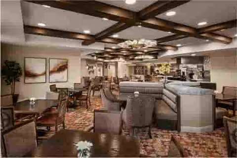 A spacious and well-lit common area with multiple tables and chairs arranged for dining or socializing. The room features a patterned carpet, wooden ceiling beams, modern light fixtures, and artwork on the walls. There is a large cushioned seating area in the center with flowers on the tables, creating a welcoming atmosphere.