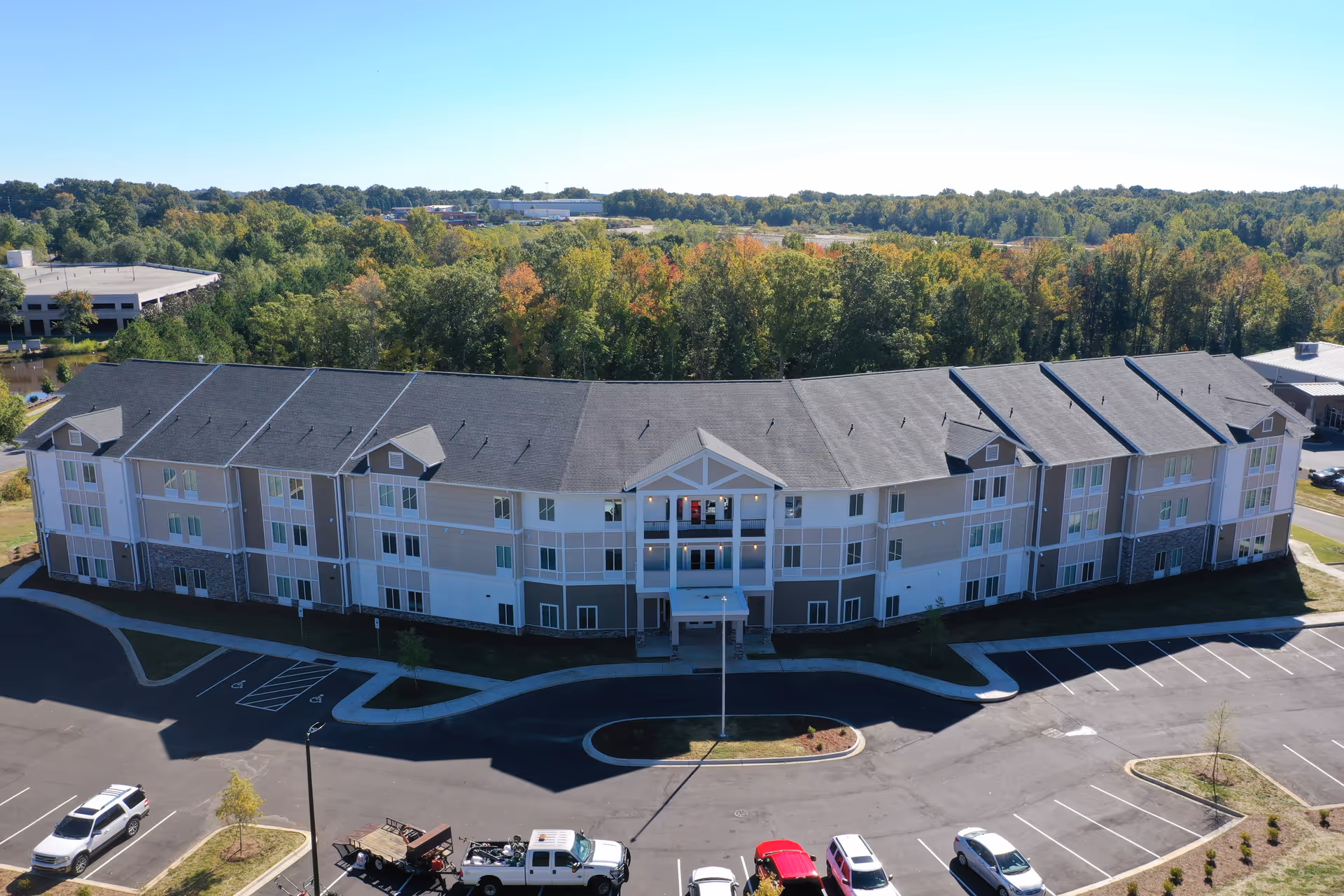 Aerial view of a large, three-story senior living facility building with a gray roof and beige exterior walls. The building is surrounded by a parking lot with several parked vehicles and is set against a backdrop of dense green trees under a clear blue sky.