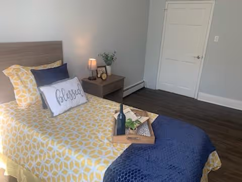 A furnished bedroom with a single bed in yellow patterned bedding and a navy throw, a nightstand with a lamp and decor, and a tray holding a wine bottle on the bed.