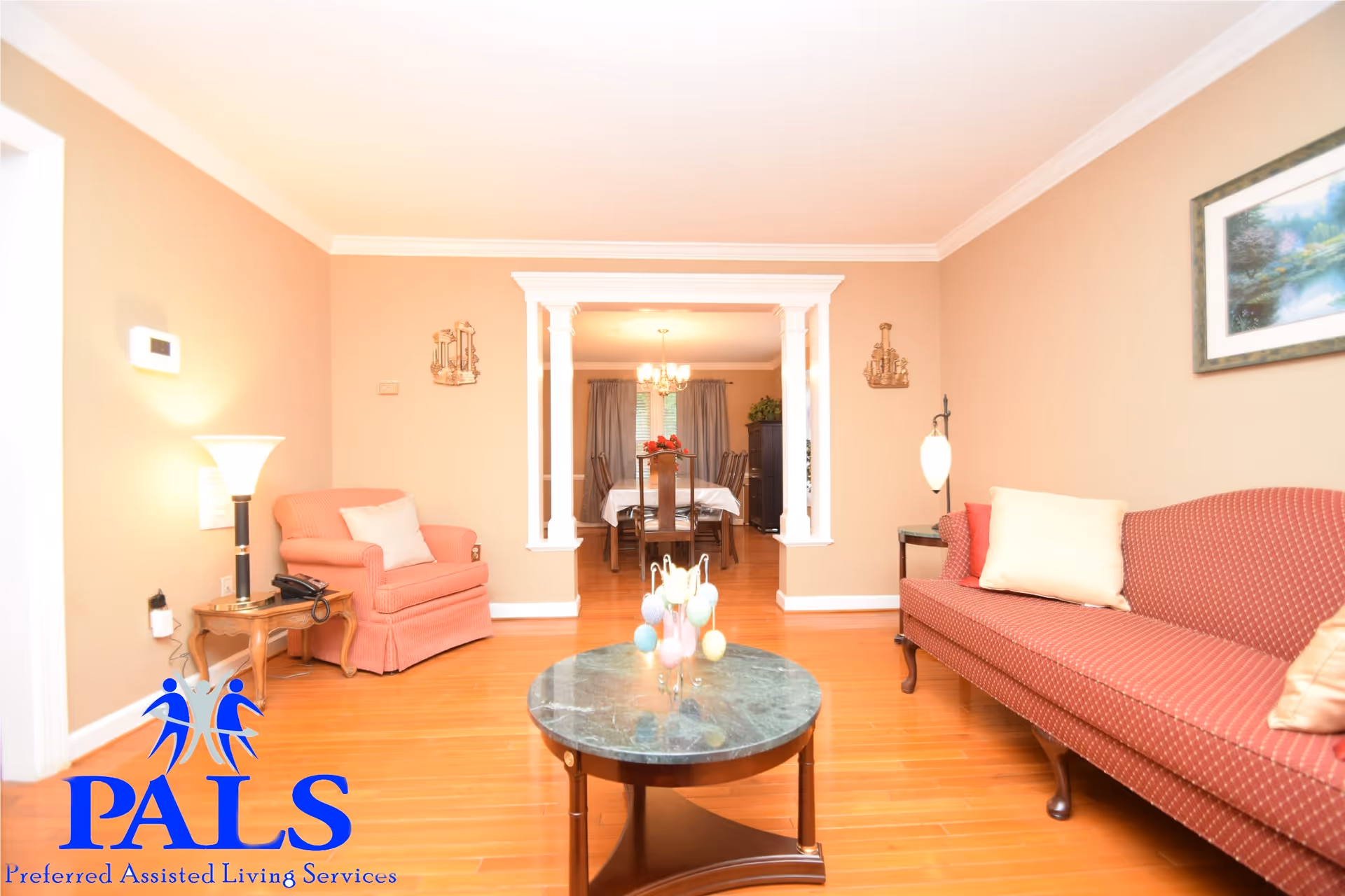 A cozy living room with wooden flooring, a red patterned sofa with cream pillows on the right, a matching armchair with a cream pillow on the left, a round marble-top coffee table in the center, and a lamp on a small wooden side table. The room opens into a dining area with a table covered with a white tablecloth and wooden chairs, illuminated by a chandelier. The walls are painted beige and decorated with framed artwork and wall sconces.