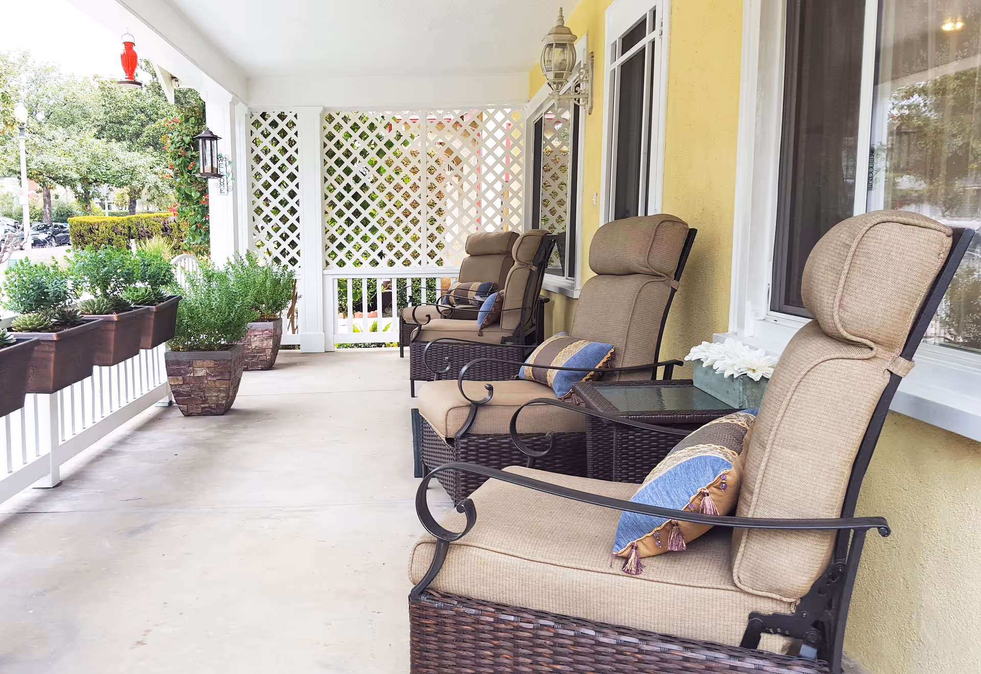A covered outdoor porch area with three cushioned wicker chairs, each with a decorative pillow. The porch has a yellow wall with windows, white railing, potted plants, and lattice panels at the far end. The setting is bright and inviting with greenery visible outside.