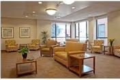 Bright communal lounge with sofas, armchairs, coffee tables, plants, framed artwork, and large windows.