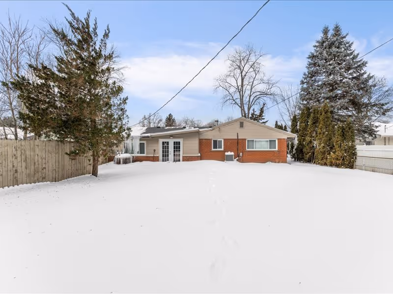 Snow-covered backyard of a single-story house with a brick and beige siding exterior, surrounded by trees and a wooden fence under a partly cloudy sky.