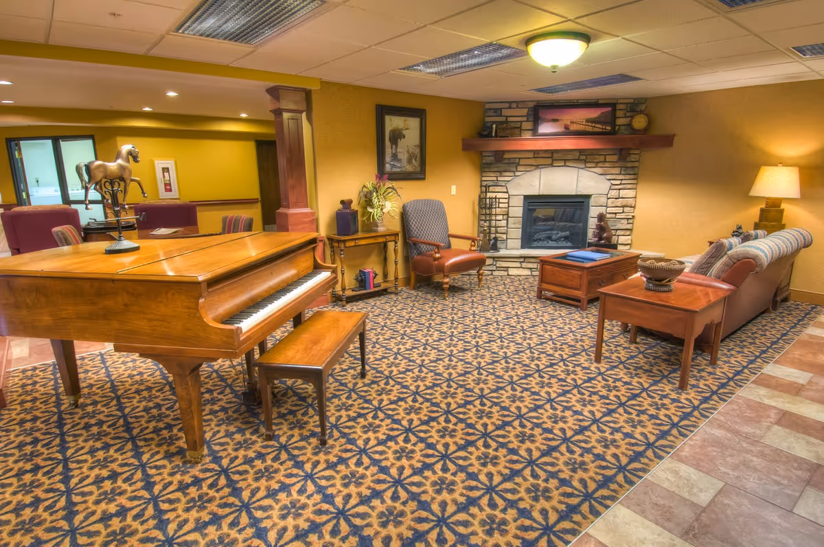 A cozy living room area with a wooden grand piano and matching bench on a patterned carpet. The room features a stone fireplace with a wooden mantel, a framed picture above it, and a comfortable seating arrangement including an armchair and a sofa with a side table and lamp. The walls are painted yellow, and there is a decorative horse sculpture on the piano.