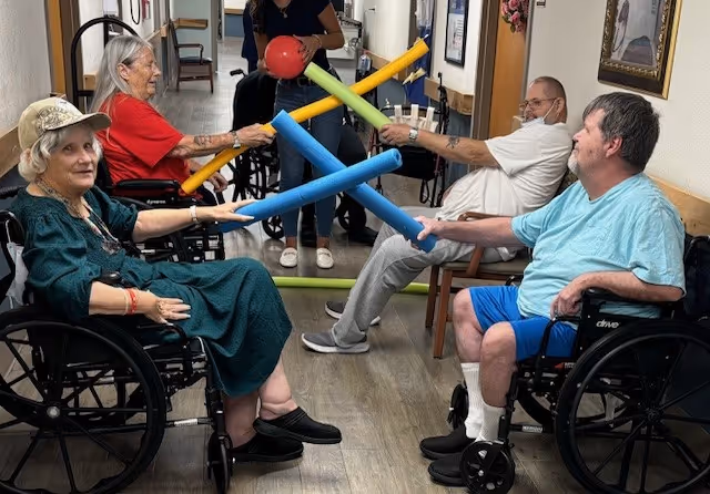 Four elderly individuals in wheelchairs are seated in a hallway, engaging in a group activity using colorful foam noodles. A staff member stands in the background overseeing the activity. The hallway has wooden flooring and framed pictures on the walls.