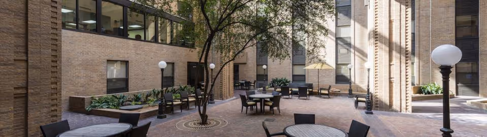 Outdoor courtyard area with brick walls surrounding it, featuring several round tables with chairs, potted plants, a tree in the center, and lamp posts with globe lights. There is also a yellow umbrella providing shade over one of the tables.