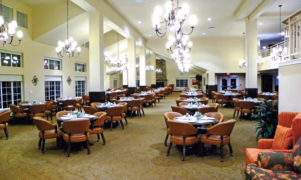 A spacious dining room with multiple round tables set with white plates, silverware, and small centerpieces. The room features high ceilings with several chandeliers, large windows along the left wall, and a carpeted floor. There are comfortable brown chairs around each table and a cozy seating area with upholstered armchairs on the right side.
