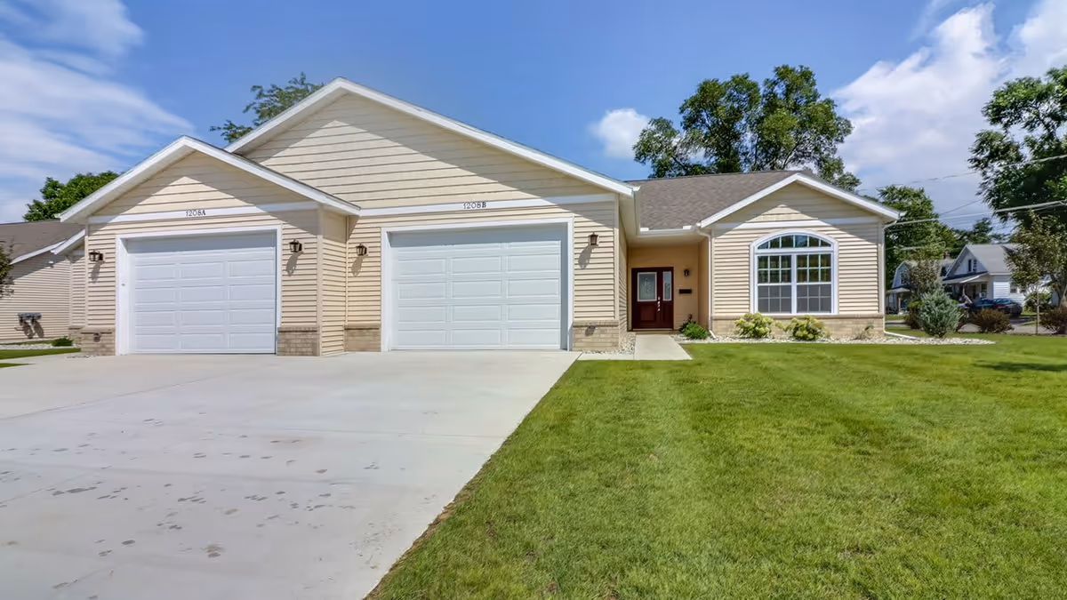 Front exterior view of a beige single-story building with two white garage doors labeled 1208A and 1208B, a red front door, a large window, and a well-maintained green lawn under a partly cloudy blue sky.