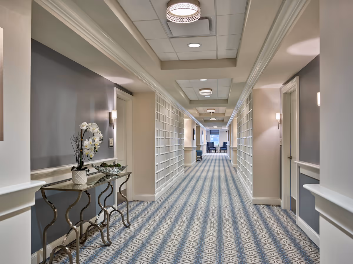 A long, well-lit hallway in an assisted living facility with patterned carpet, light gray and beige walls, decorative wall panels, and a glass table with a white orchid and decorative bowl. At the far end, there are two blue chairs near a window.