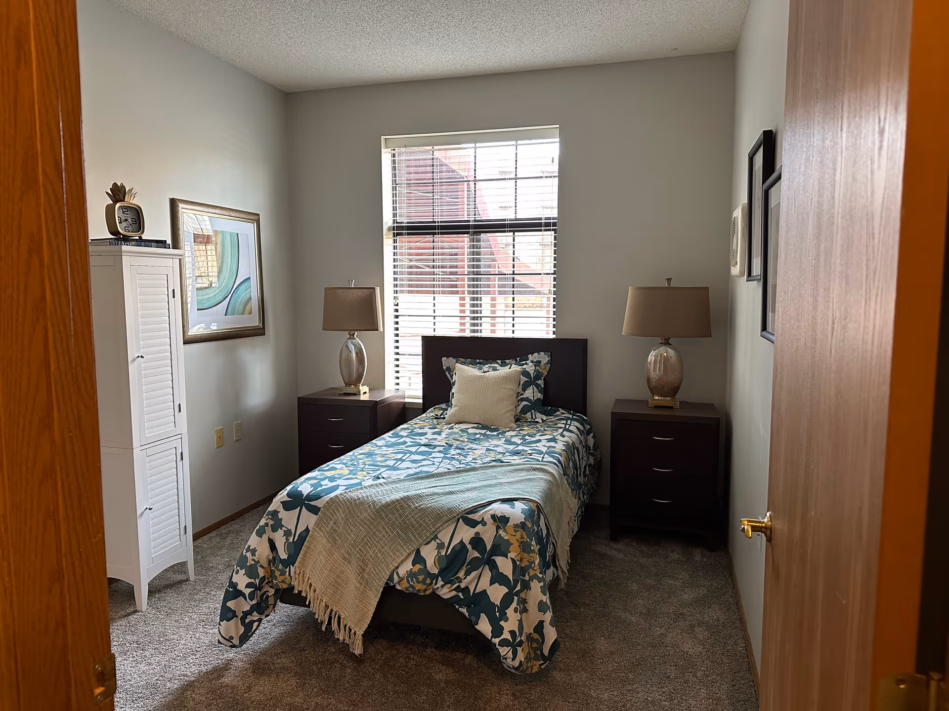 A small bedroom with a single bed covered in a floral patterned bedspread and a beige throw blanket. There are two dark wooden nightstands on either side of the bed, each with a lamp. A window with blinds is centered on the back wall, letting in natural light. On the left side of the room, there is a white cabinet with a clock and some books on top, and a framed abstract painting hangs on the wall above it. The room has beige carpet and light-colored walls.